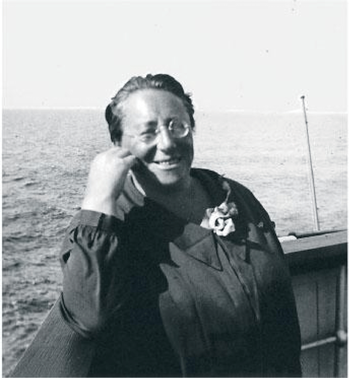 Cheerful Emmy on a boat, wearing dark clothing with a pale flower in her collar, round glasses and a smile.